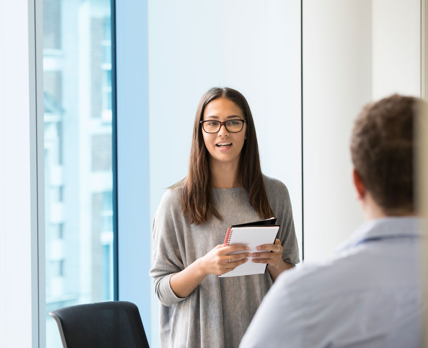 A woman stands near a big window holding a notebook and addressing a room of co-workers